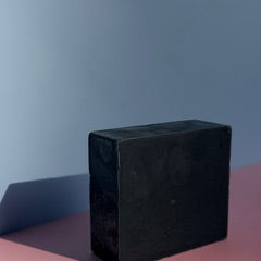 Wrapped bar of charcoal soap with grapes, oil in a beaker, a glass jar with cocoa butter wafers, and plant cuttings on a whitewashed wood background.