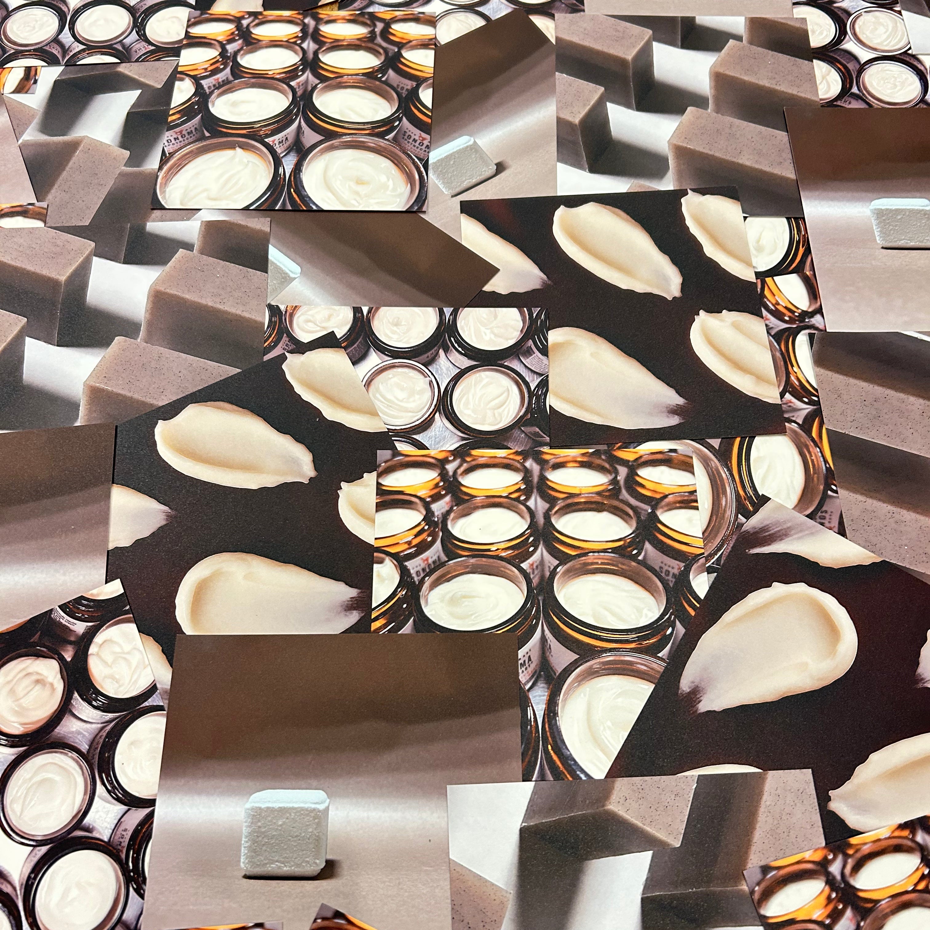 Wrapped bar of charcoal soap with grapes, oil in a beaker, a glass jar with cocoa butter wafers, and plant cuttings on a whitewashed wood background.