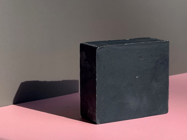 Wrapped bar of charcoal soap with grapes, oil in a beaker, a glass jar with cocoa butter wafers, and plant cuttings on a whitewashed wood background.