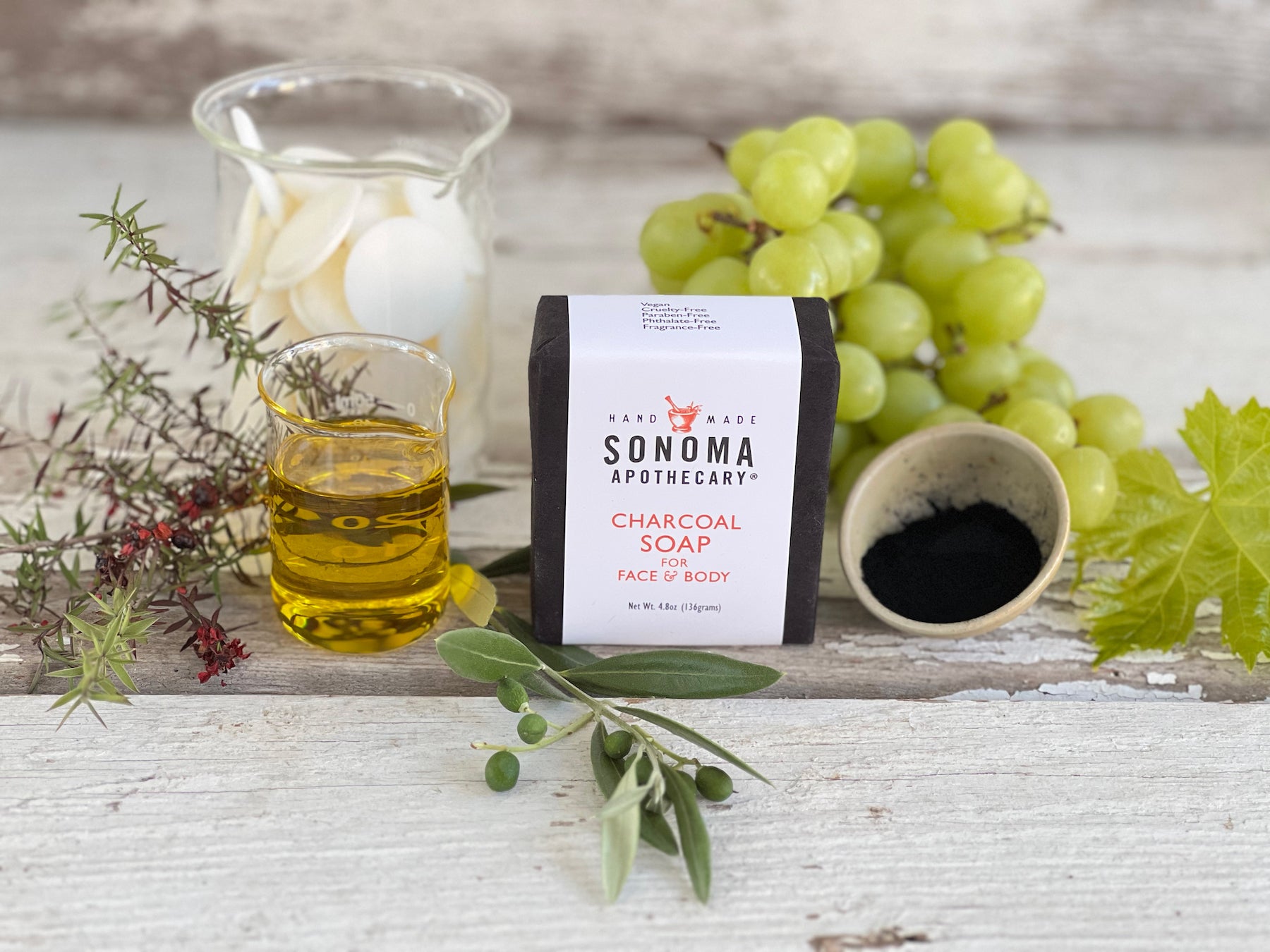 Wrapped bar of charcoal soap with grapes, oil in a beaker, a glass jar with cocoa butter wafers, and plant cuttings on a whitewashed wood background.
