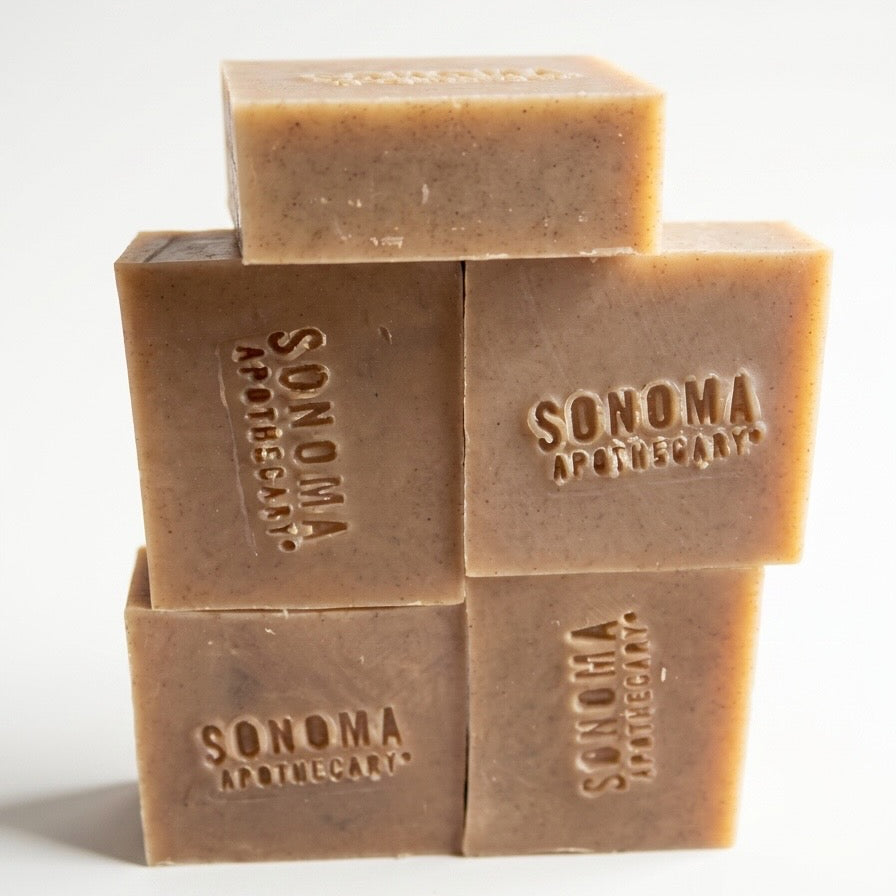 Collection of body scrub soap bars with plants, grapes, a sunflower, cut fruit, natural bristle brush against a whitewashed wood structure.