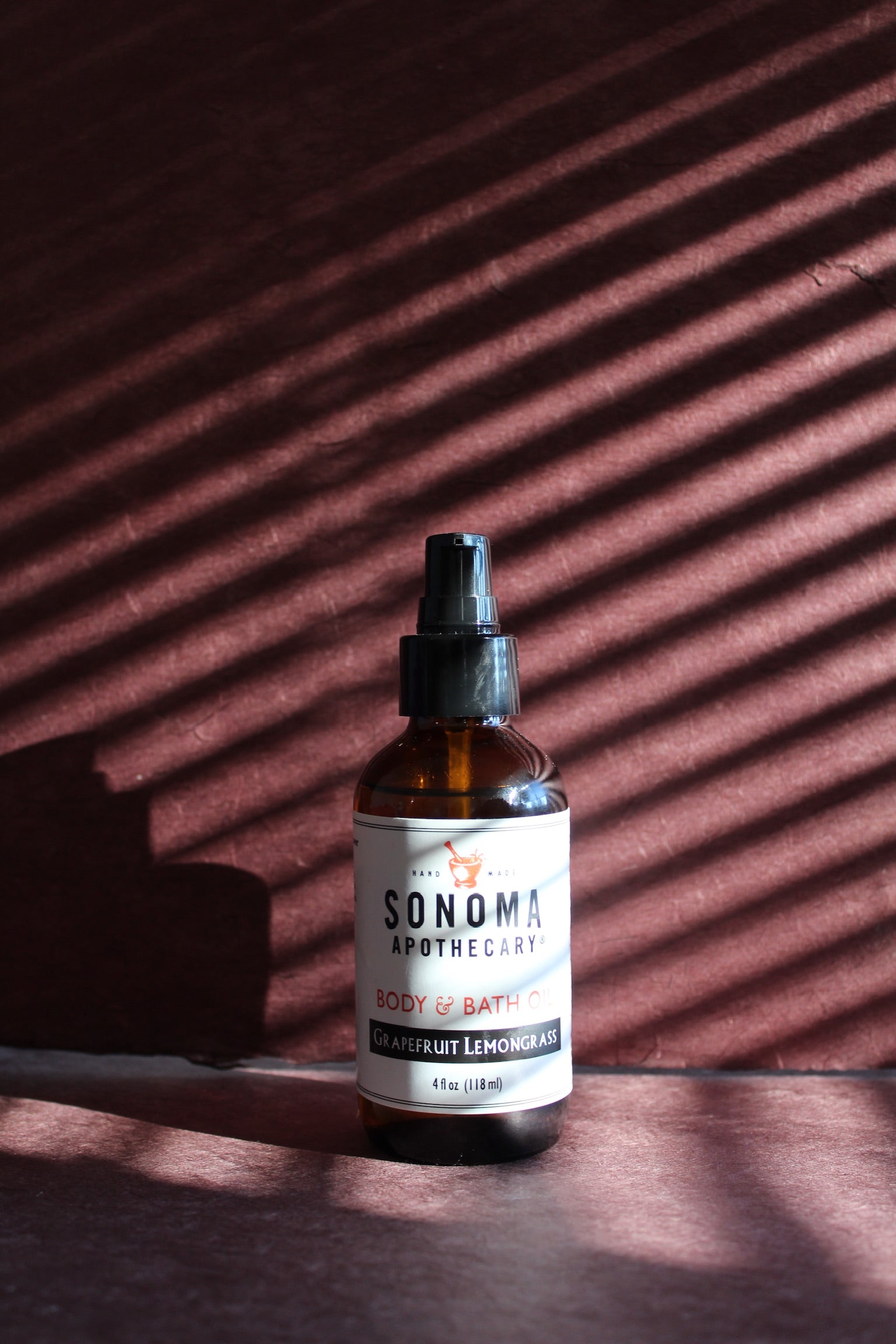 Collection of different varieties of body oils in two different sized glass bottles with plants, cut fruit and grapes on a whitewashed bench against a white wooden background..