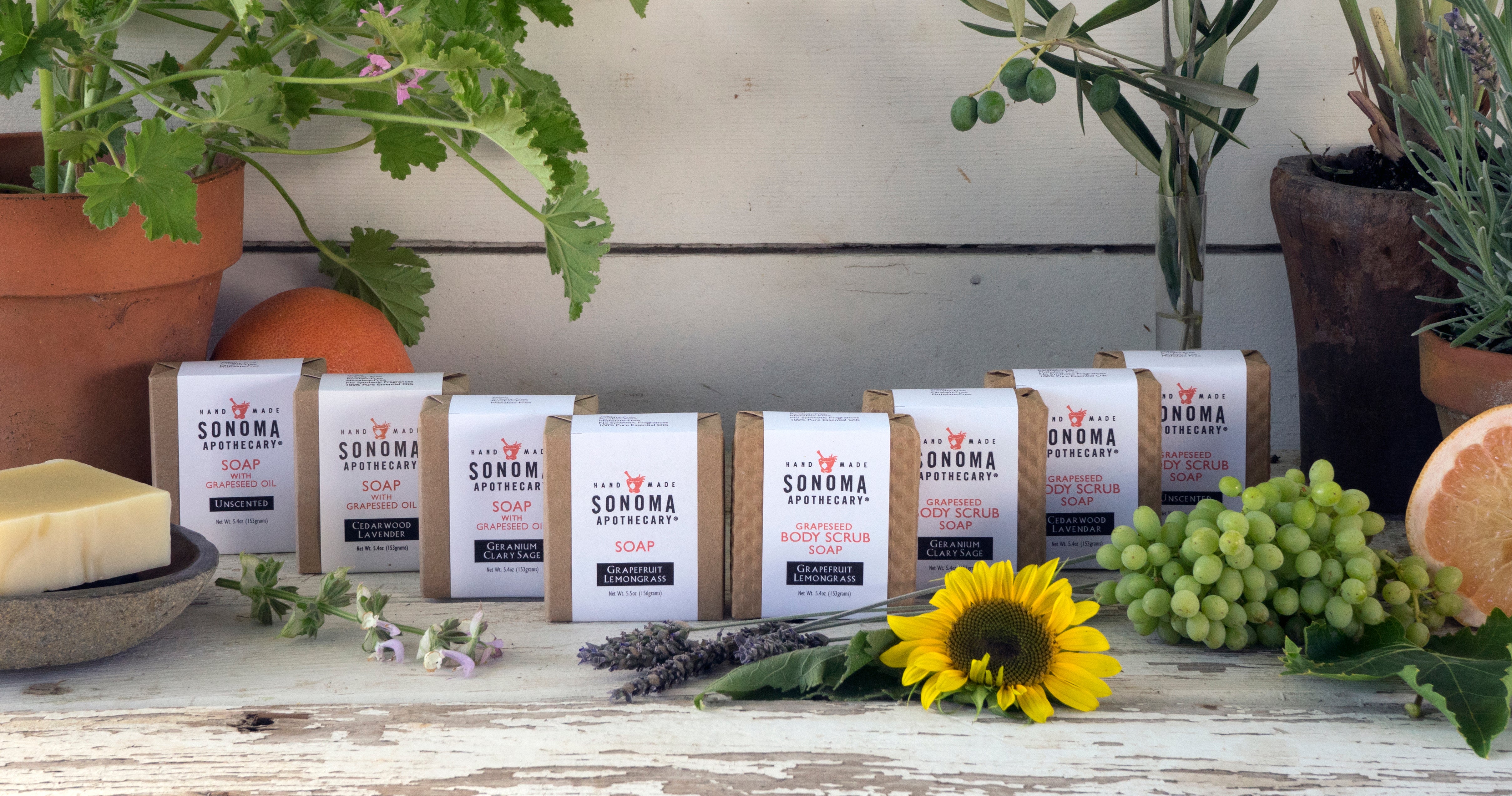 Collection of Natural bar soaps with grapes, plants, cut fruit, and sunflower with whitewashed wooden structure in background.