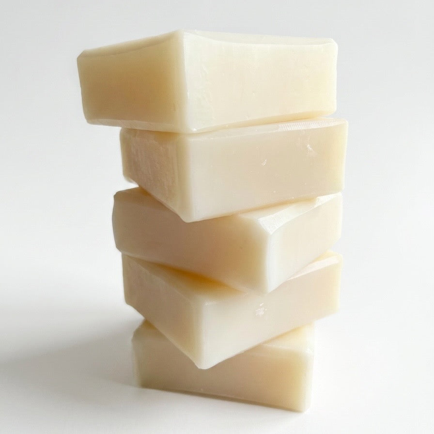 Collection of Natural bar soaps with grapes, plants, cut fruit, and sunflower with whitewashed wooden structure in background.