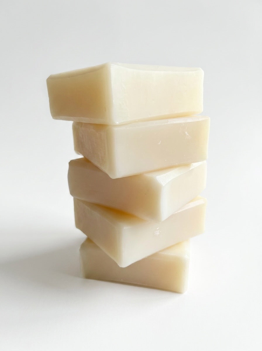 Collection of Natural bar soaps with grapes, plants, cut fruit, and sunflower with whitewashed wooden structure in background.