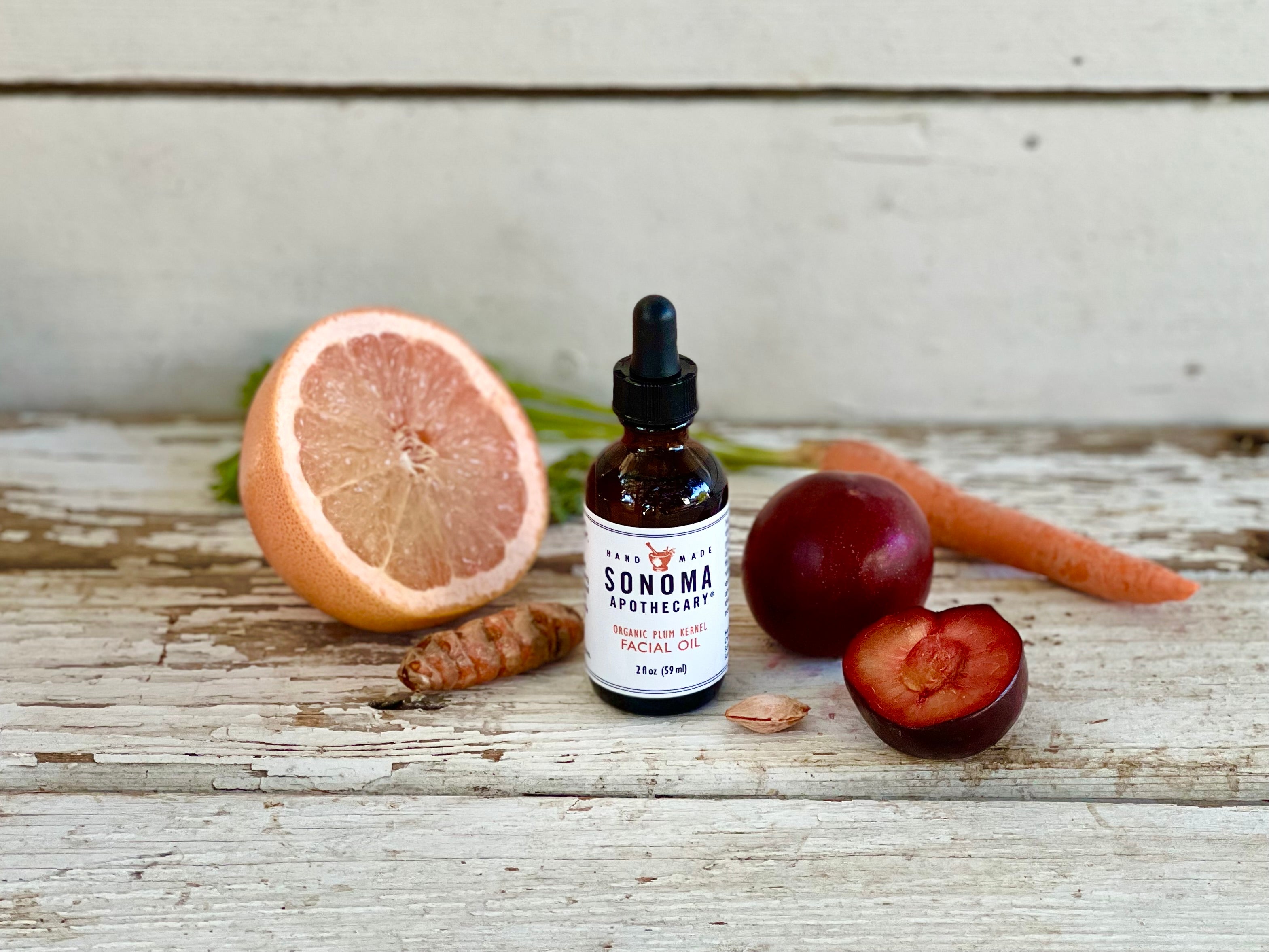 dark glass bottle with white label and dropper containing oil with cut fruit, and a carrot on a whitewashed wooden board.