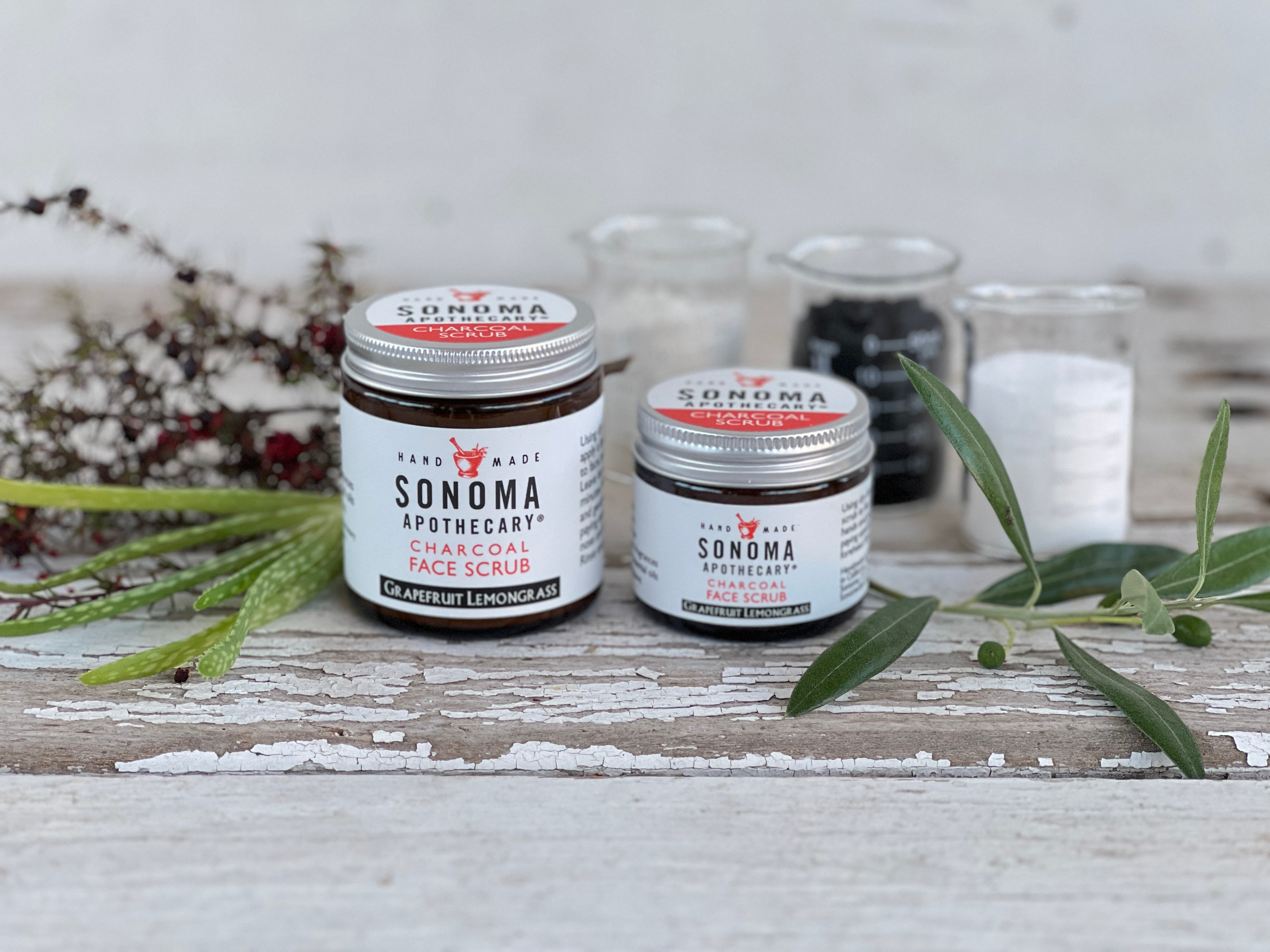 Two different sized glass jars with white labels; three glass beakers with activated charcoal, natural clay, jojoba wax beads; with plant cuttings displayed on a white wood board.