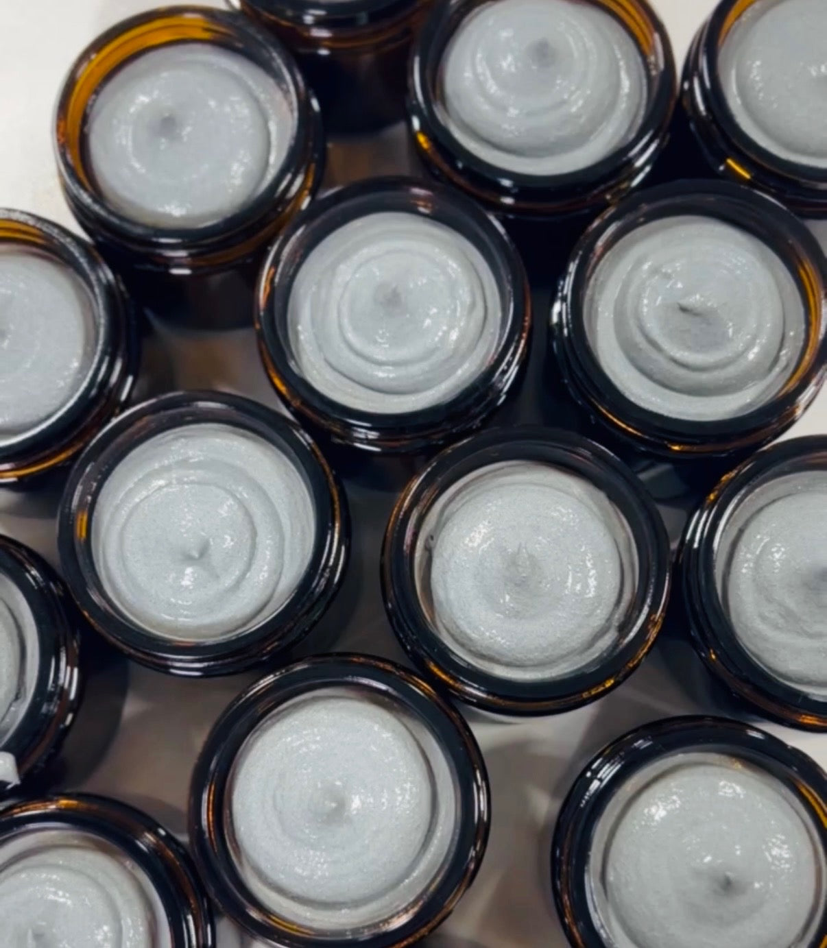 Two different sized glass jars with white labels; three glass beakers with activated charcoal, natural clay, jojoba wax beads; with plant cuttings displayed on a white wood board.