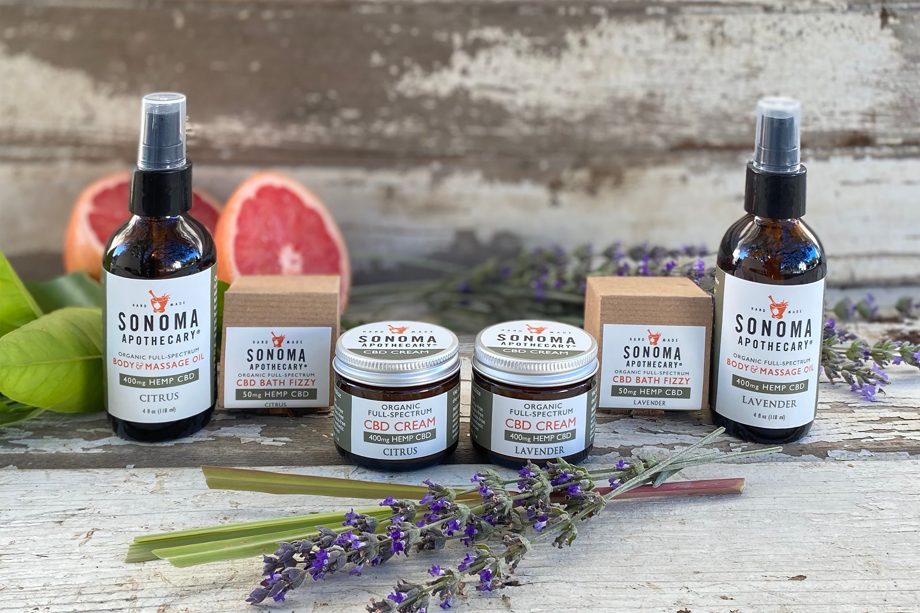 Collection of CBD products packaged in different glass bottles and boxes with fresh plant cuttings and cut fruit against a whitewashed wood background.