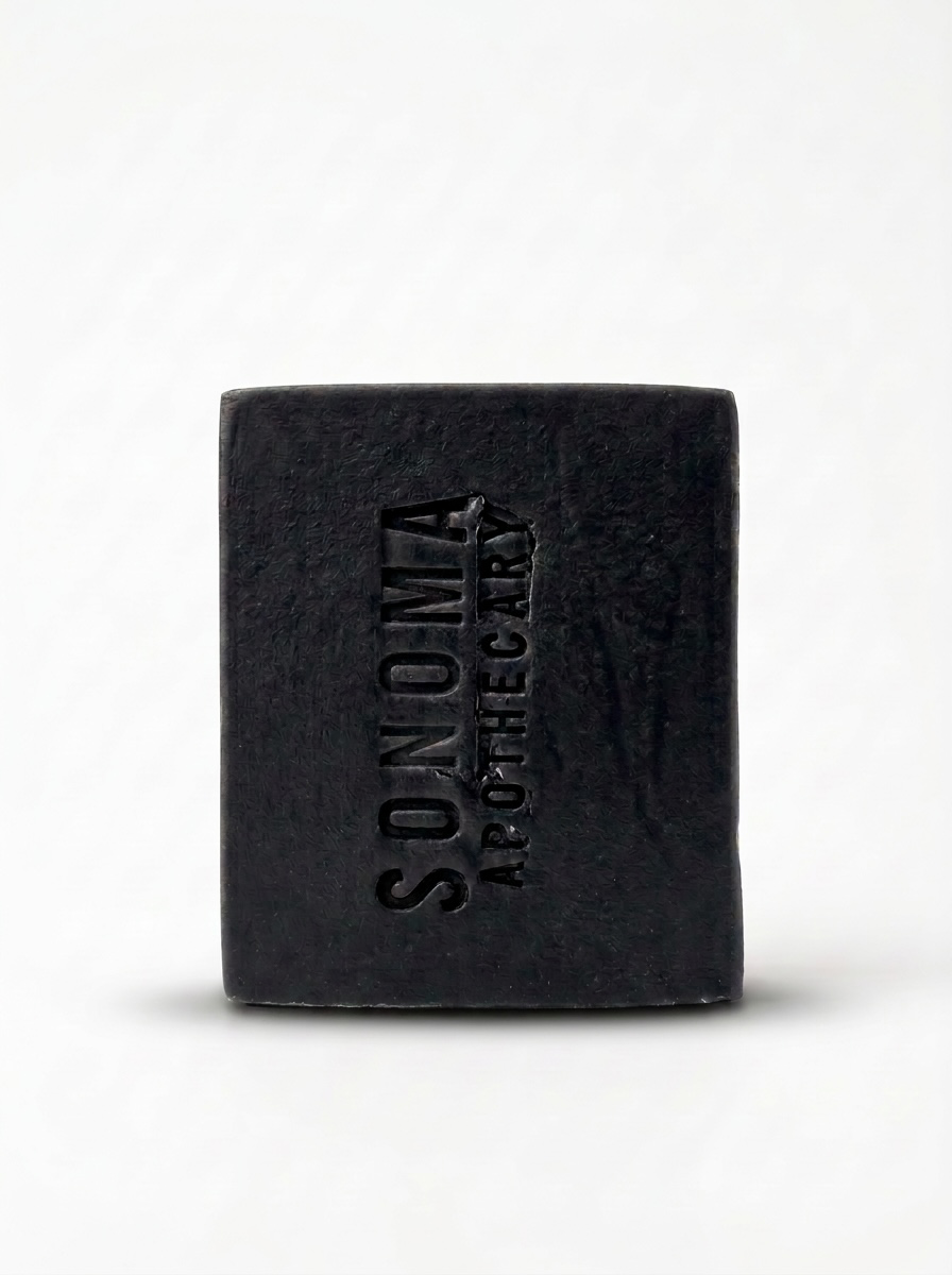 Wrapped bar of charcoal soap with grapes, oil in a beaker, a glass jar with cocoa butter wafers, and plant cuttings on a whitewashed wood background.