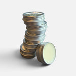 A stack of round silver metal lip balm tins with one tin lying on its side, showing the pale yellow balm inside. 