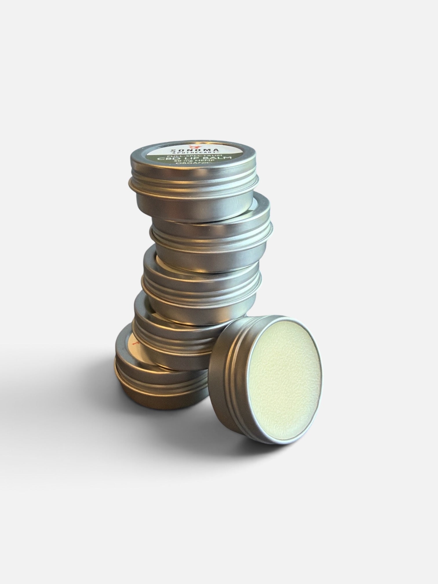 A stack of round silver metal lip balm tins with one tin lying on its side, showing the pale yellow balm inside. 